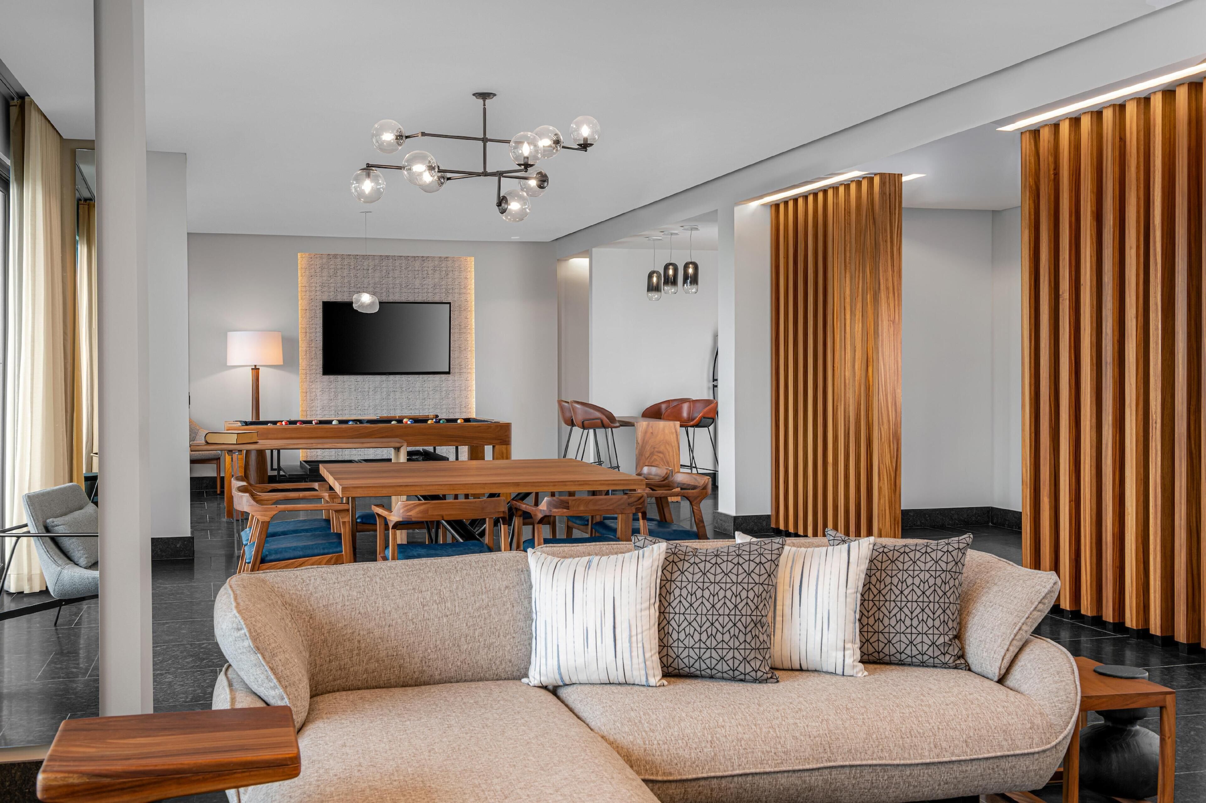 Ein modernes Wohnzimmer mit einer Eckcouch, einem Holzessttisch und Stühlen. Ein Flachbildfernseher ist an der Wand montiert, und über dem Essbereich hängt eine Kristallleuchter.