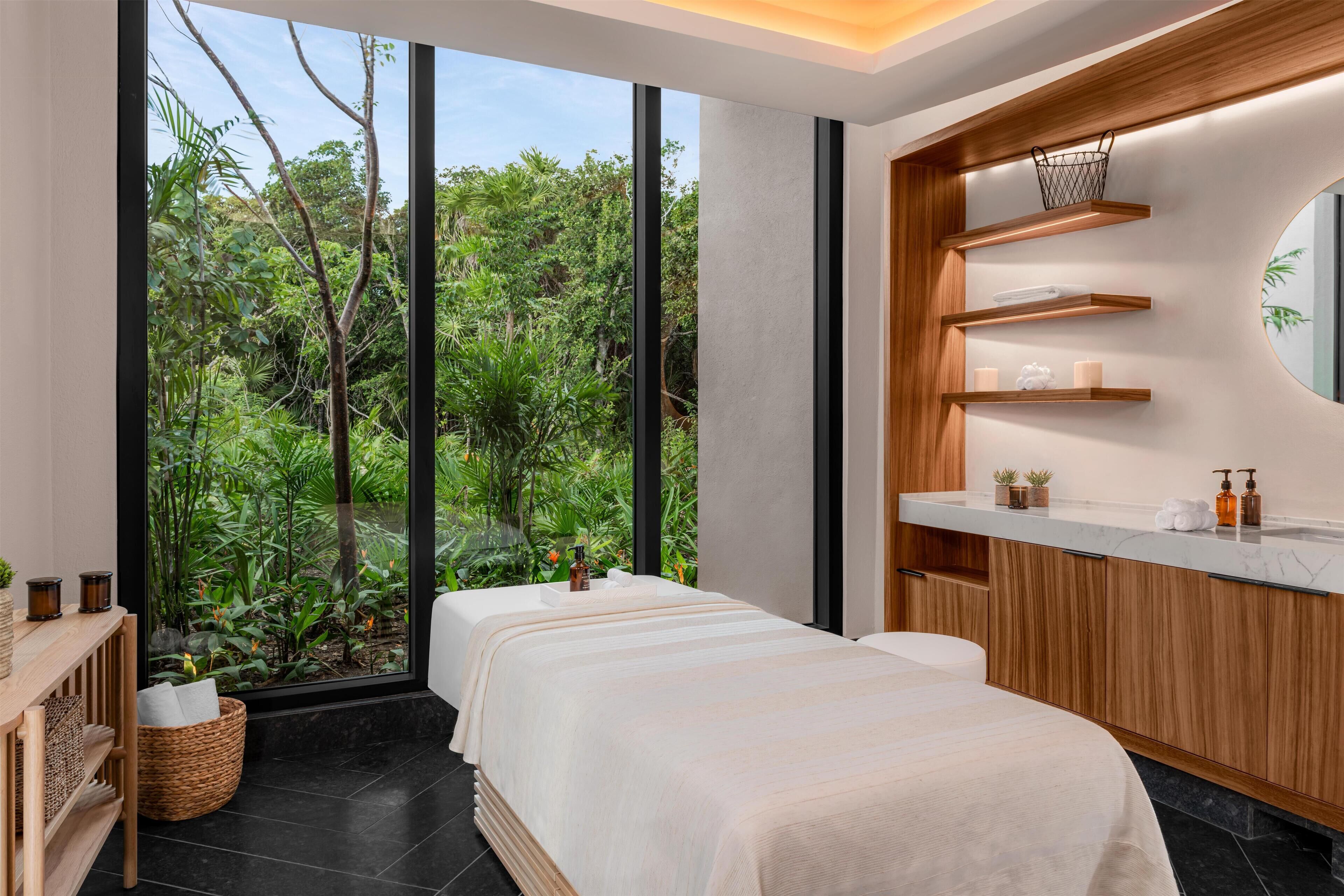 Un'elegante sala spa con una grande finestra vista su un rigoglioso giardino, un settimino in legno con ripiani e un lettino da massaggio.