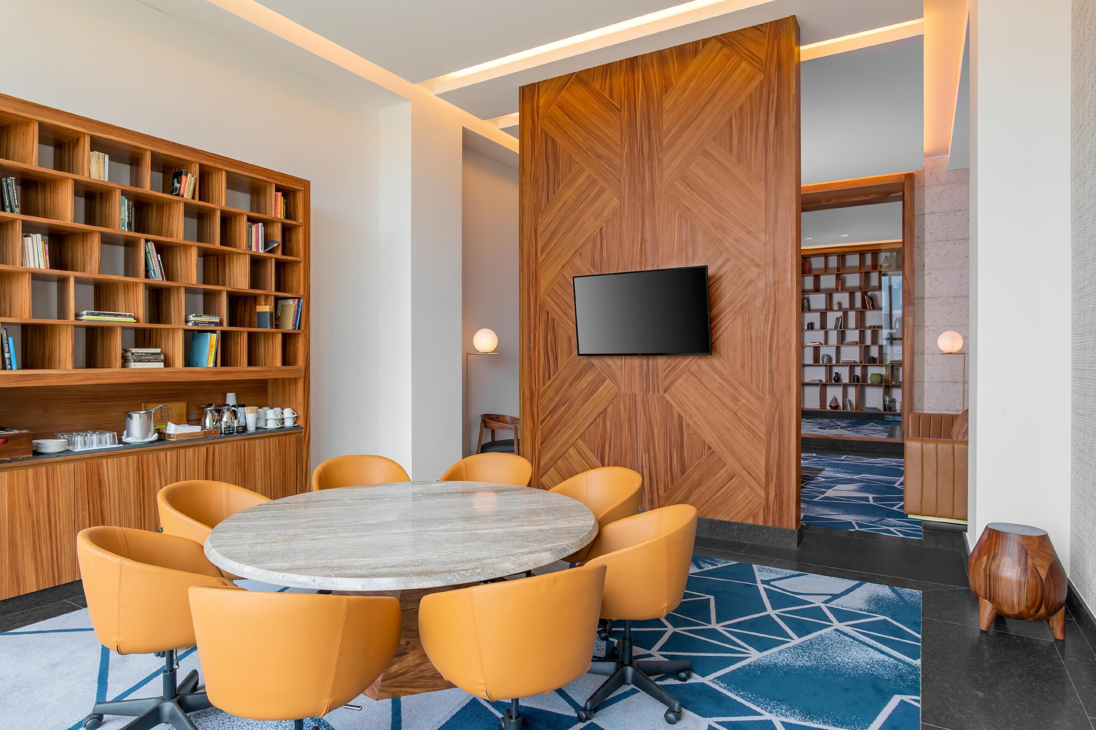 Een moderne vergaderruimte met een ronde tafel, oranje stoelen, een boekenkast, een flatscreen televisie en een kamerbreed tapijt met dessin.