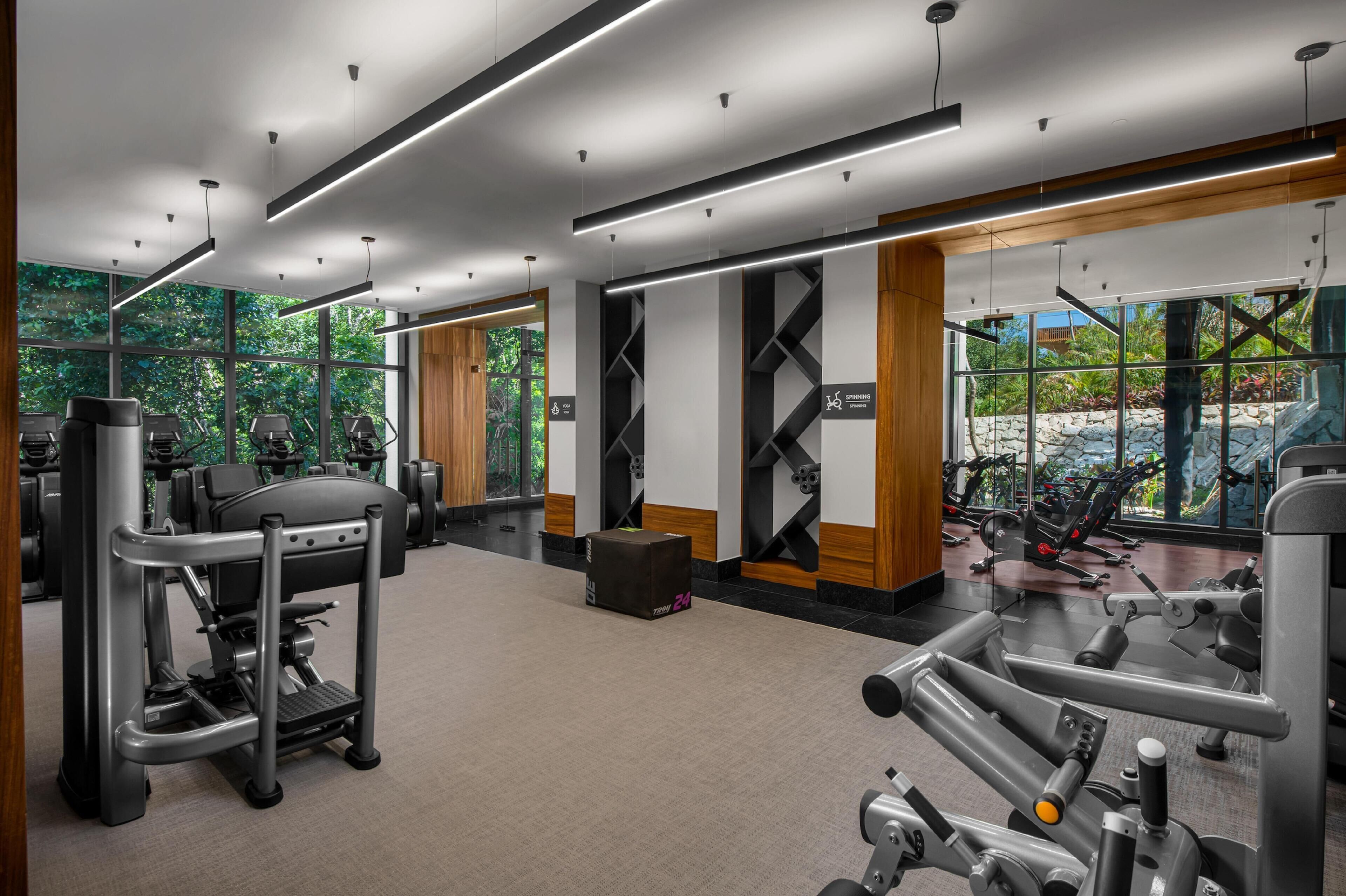 A modern gym with various exercise equipment, large windows offering views of greenery, and a clean, well-lit interior.