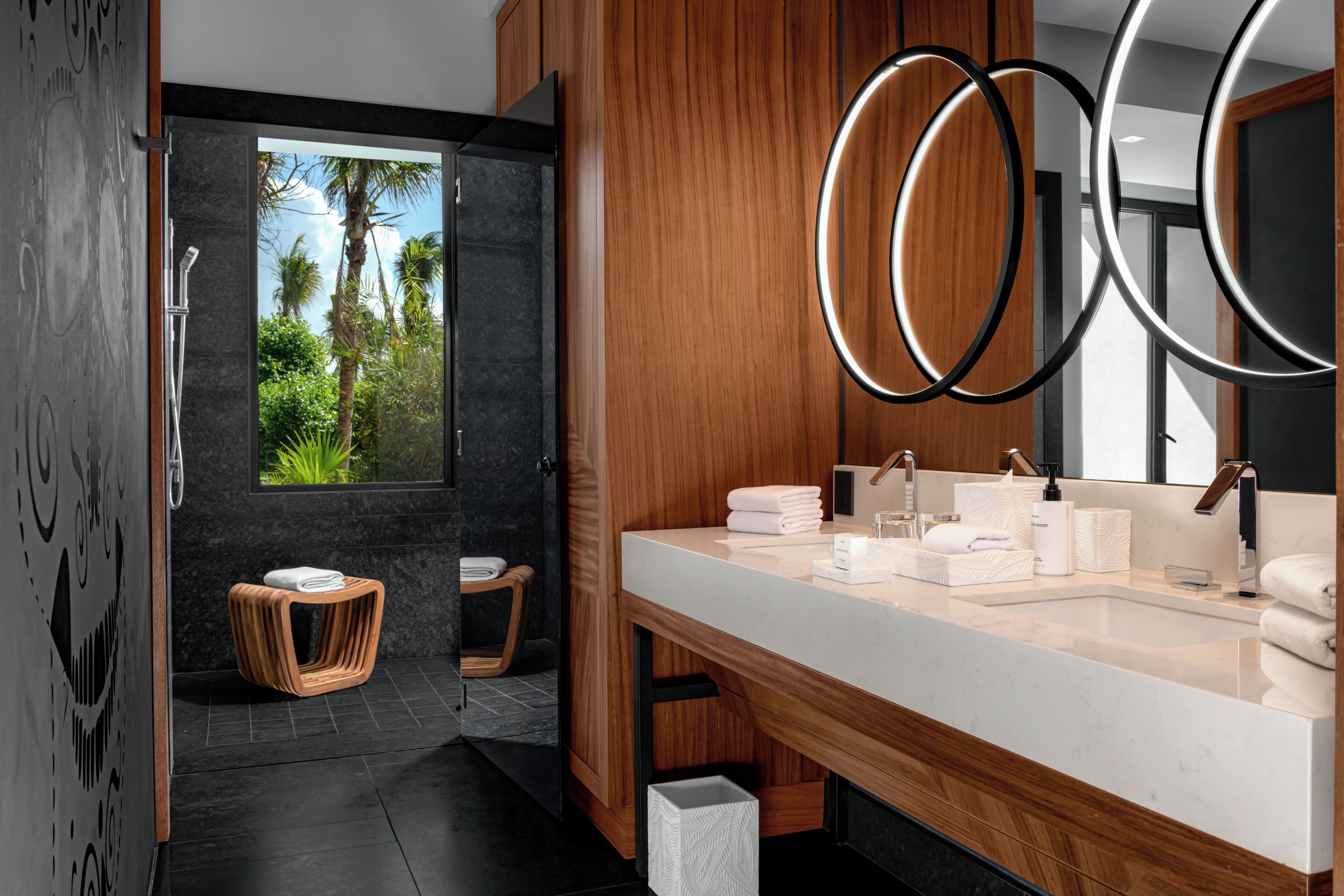 Un baño moderno con un espejo grande, armarios de madera, lavamanos de mármol y vistas a palmeras.