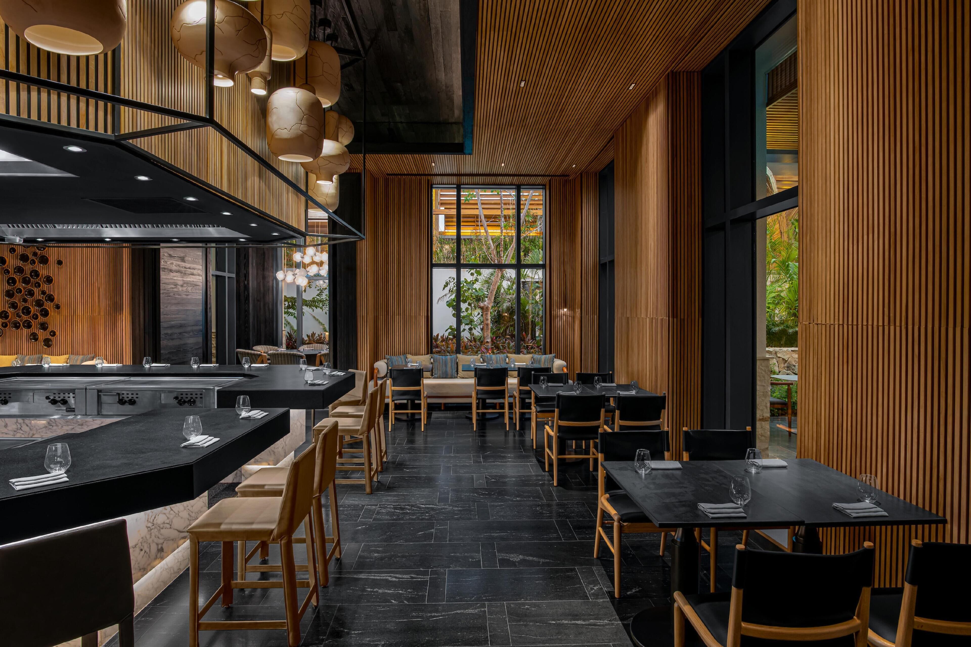 Un restaurante moderno con interior en tonos negro y madera, que presenta un área central de comedor con mesas y sillas, una zona de bar con mostrador, y ventanas amplias con vistas al exterior.