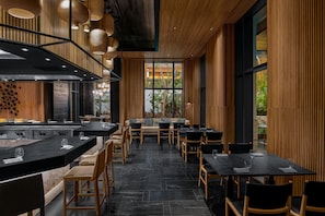 A modern restaurant with a black and wood interior, featuring a central dining area with tables and chairs, a bar area with a counter, and large windows providing views of the outdoors.