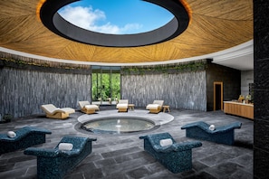A modern spa area with a central hot tub, lounge chairs, and a view of greenery through large windows.