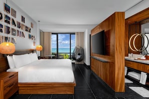 A modern hotel room with a large bed, a TV, wooden paneling, a balcony with a view of the ocean, and artwork on the walls.