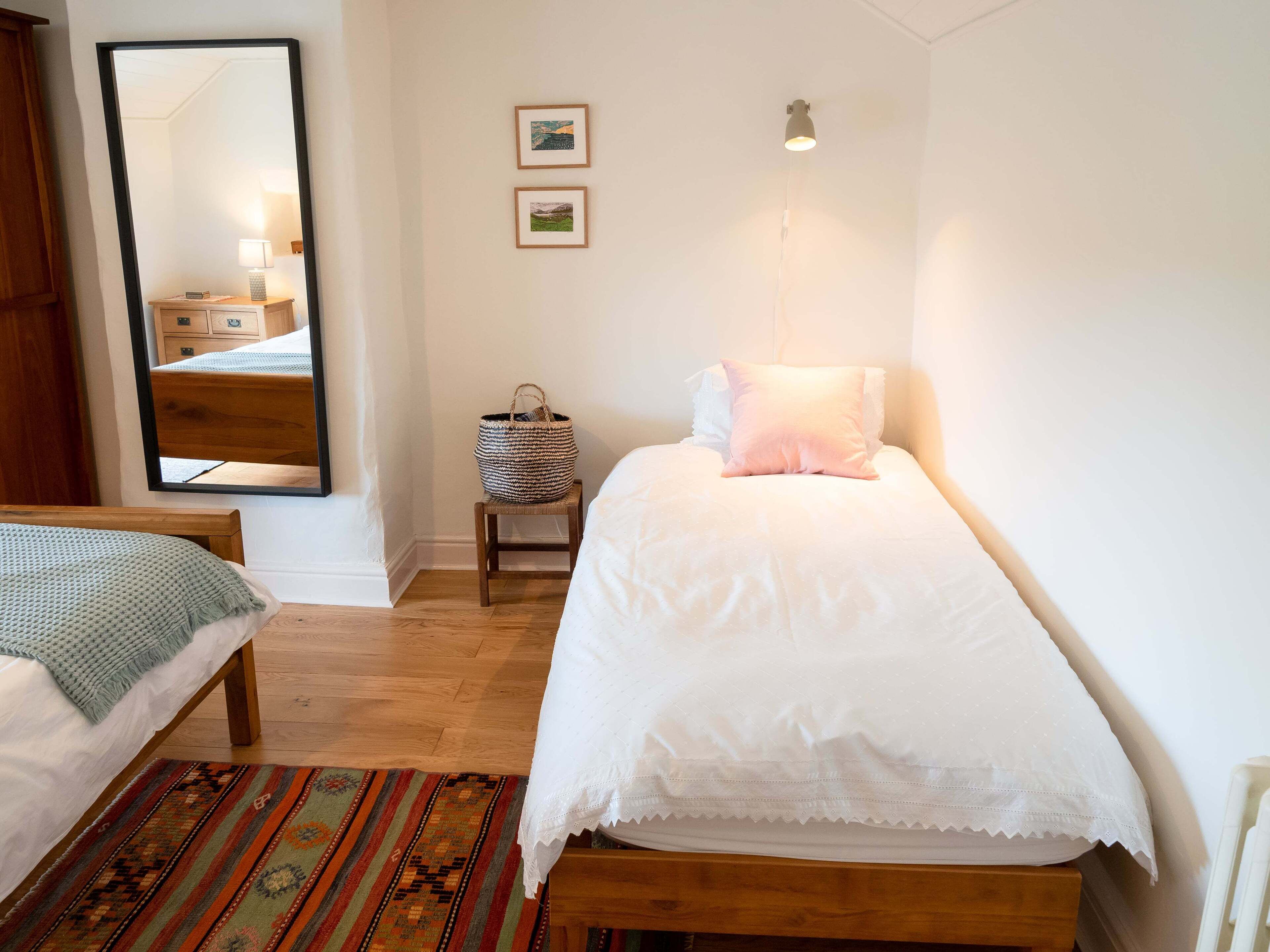 Un dormitorio con una cama, un banco, una mesita con una cesta, un espejo y dos cuadros en la pared.