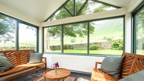Uma sala de sol com móveis de madeira, um tapete listrado e vista para uma paisagem verde através de grandes janelas.