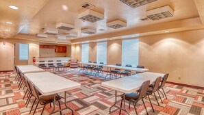 A conference room with multiple tables and chairs, a patterned carpet, and a kitchen area in the background. - Express Inn & Suites (Norman)