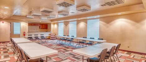 A conference room with multiple tables and chairs, a patterned carpet, and a kitchen area in the background.