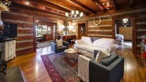 A log cabin interior with a wooden ceiling, a chandelier, a white sofa, a patterned rug, and a television.