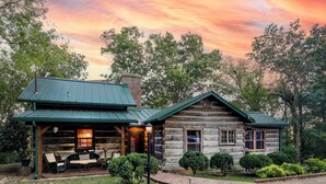 A rustic log cabin with a covered porch and a fireplace, surrounded by lush greenery.