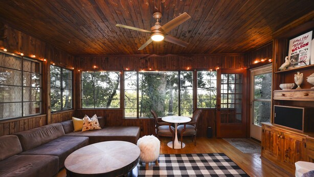 A cozy cabin interior with a large wooden ceiling, a ceiling fan, a checkered floor, a sofa, a round table, and a dining table with chairs.