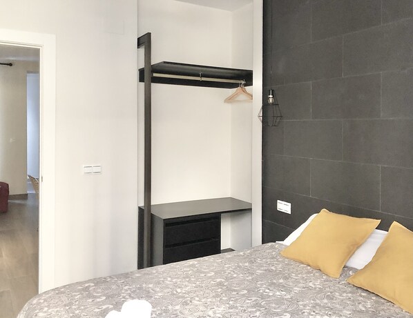 Un dormitorio con cama, mesita de noche, perchero y pared revestida de baldosas oscuras.