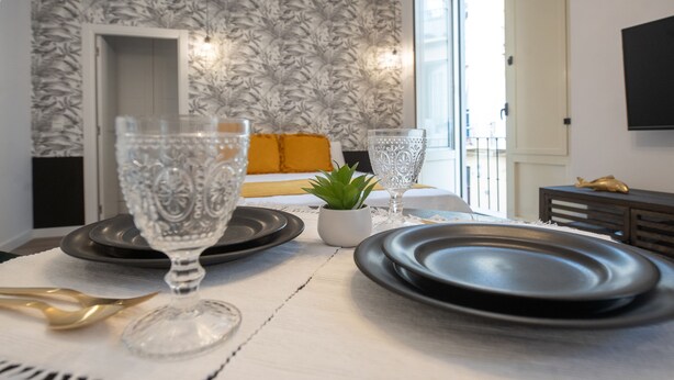 Una mesa de comedor dispuesta con platos, vasos y un tenedor con mango dorado. Hay una pequeña planta en maceta sobre la mesa.