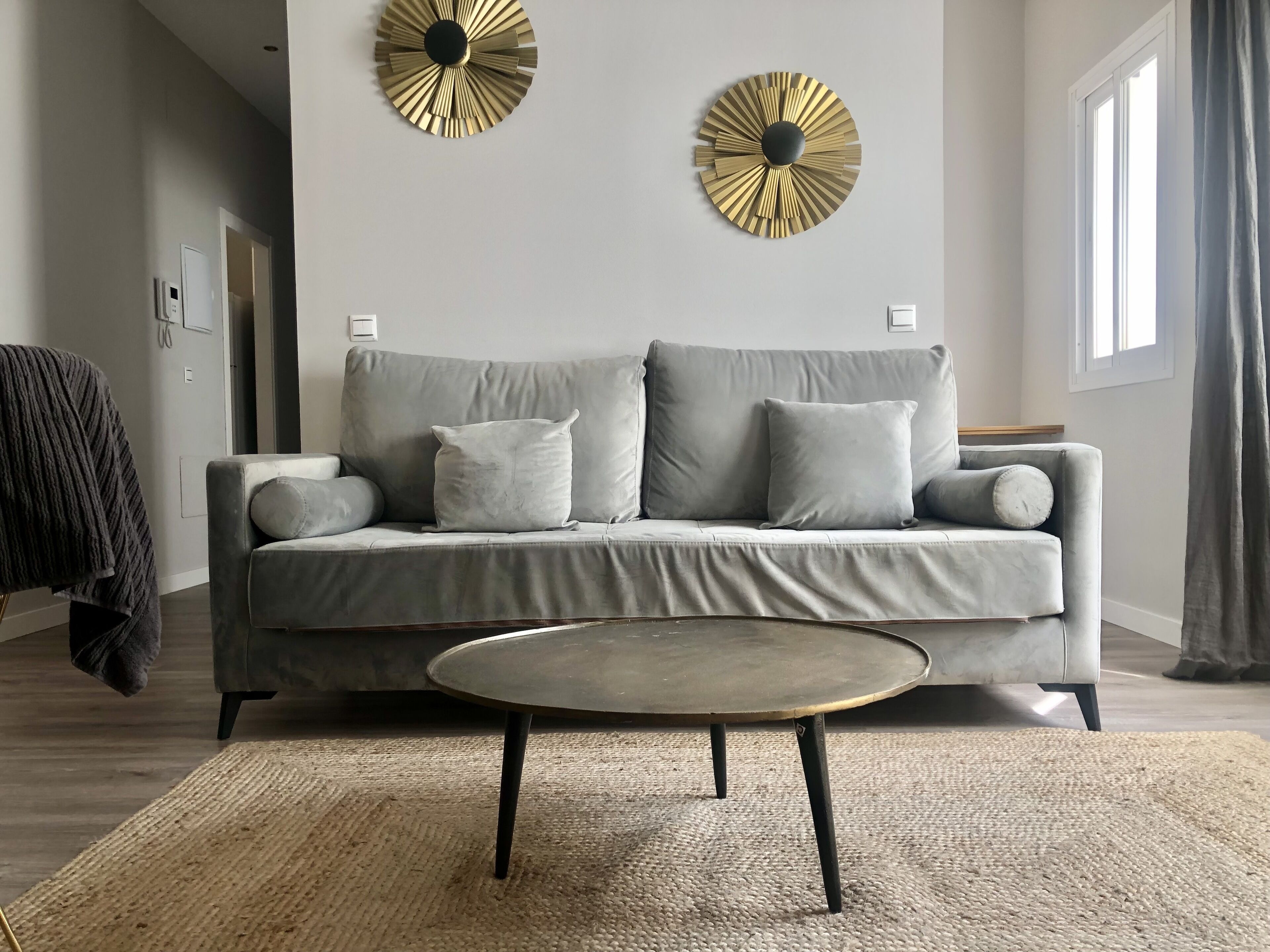 Una sala de estar moderna con un sofá gris, dos detalles decorativos dorados en la pared, una mesa de centro redonda y un ventanal con cortinas.