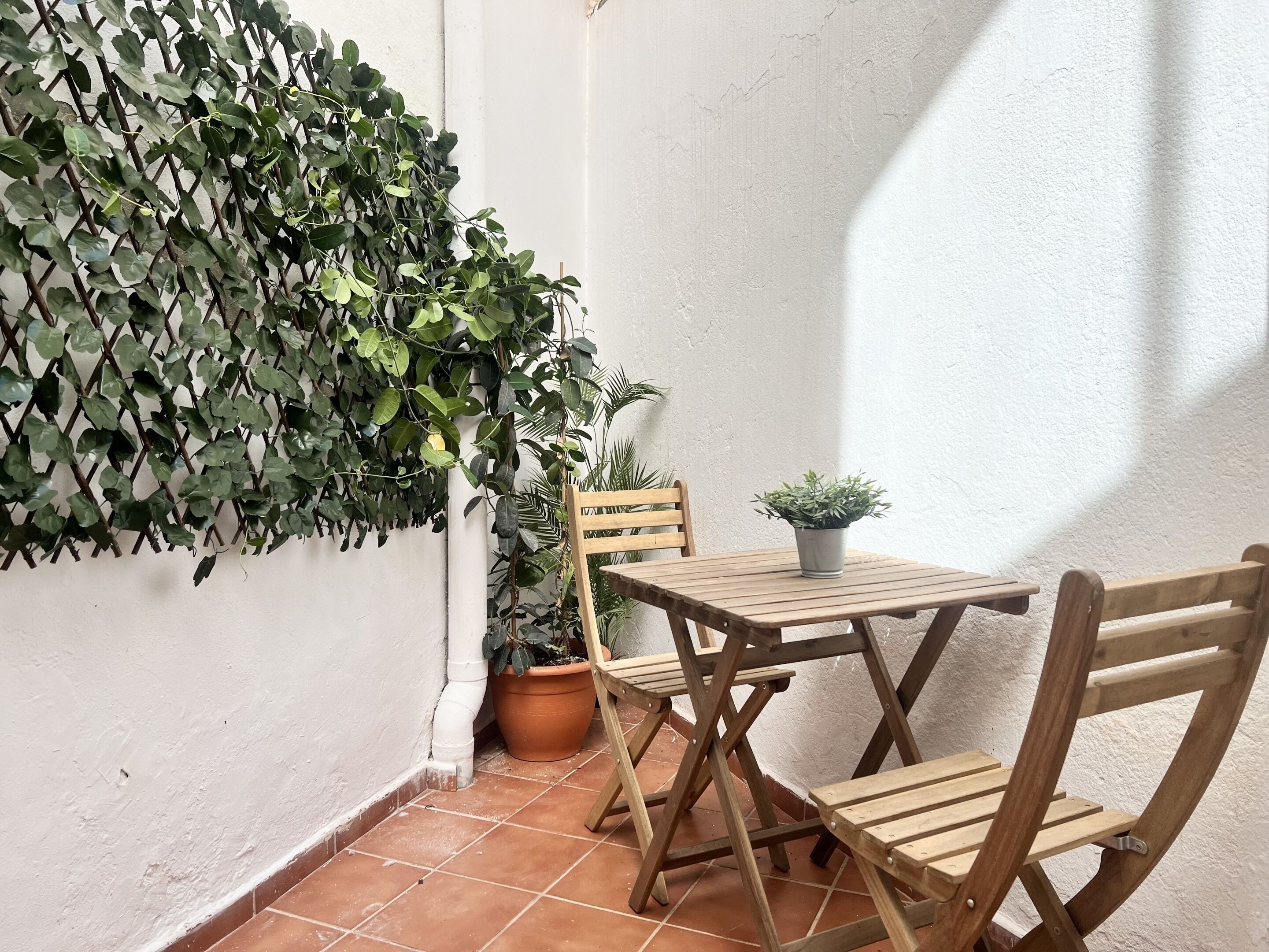 Un pequeño patio al aire libre con una mesa y sillas de madera, rodeado de plantas en macetas y una enredadera.