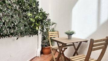 Un pequeño patio al aire libre con una mesa y sillas de madera, rodeado de plantas en macetas y una enredadera.