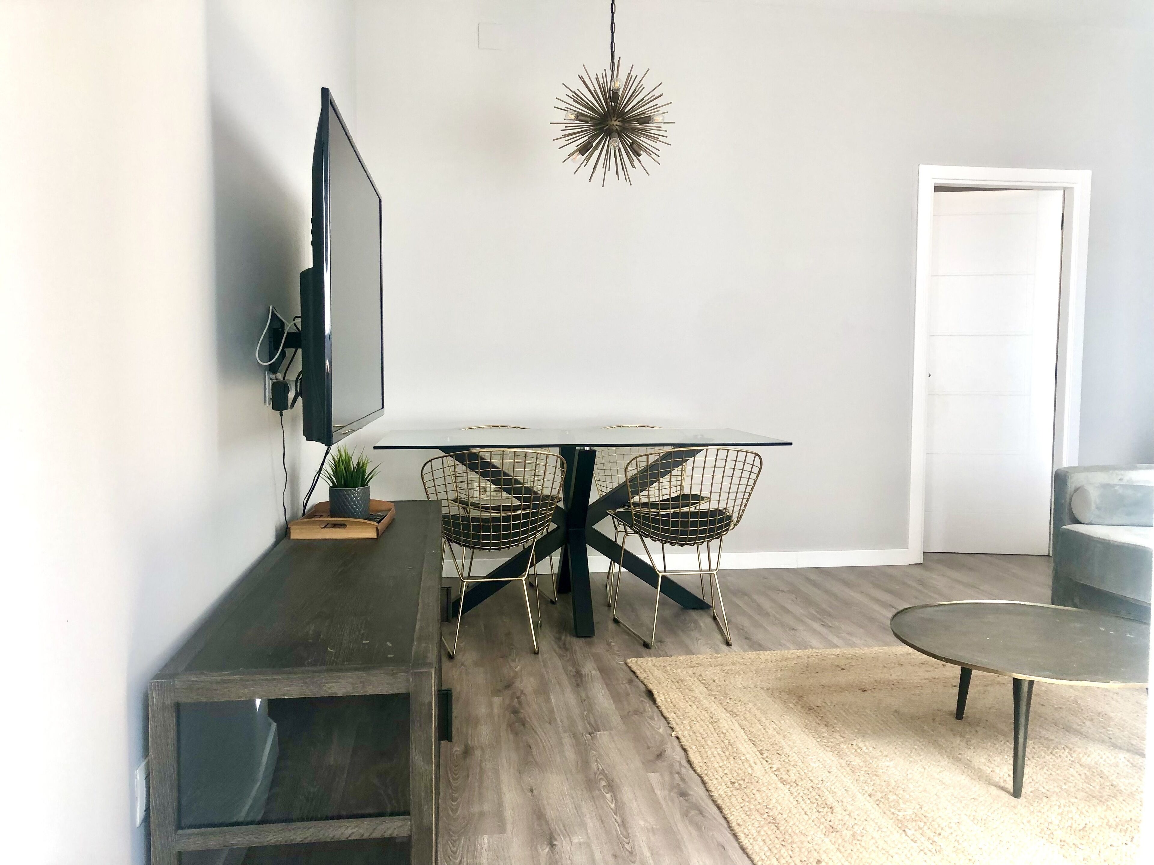 A modern living room with a flat-screen TV, a round dining table with wire chairs, a small side table, and a decorative light fixture.