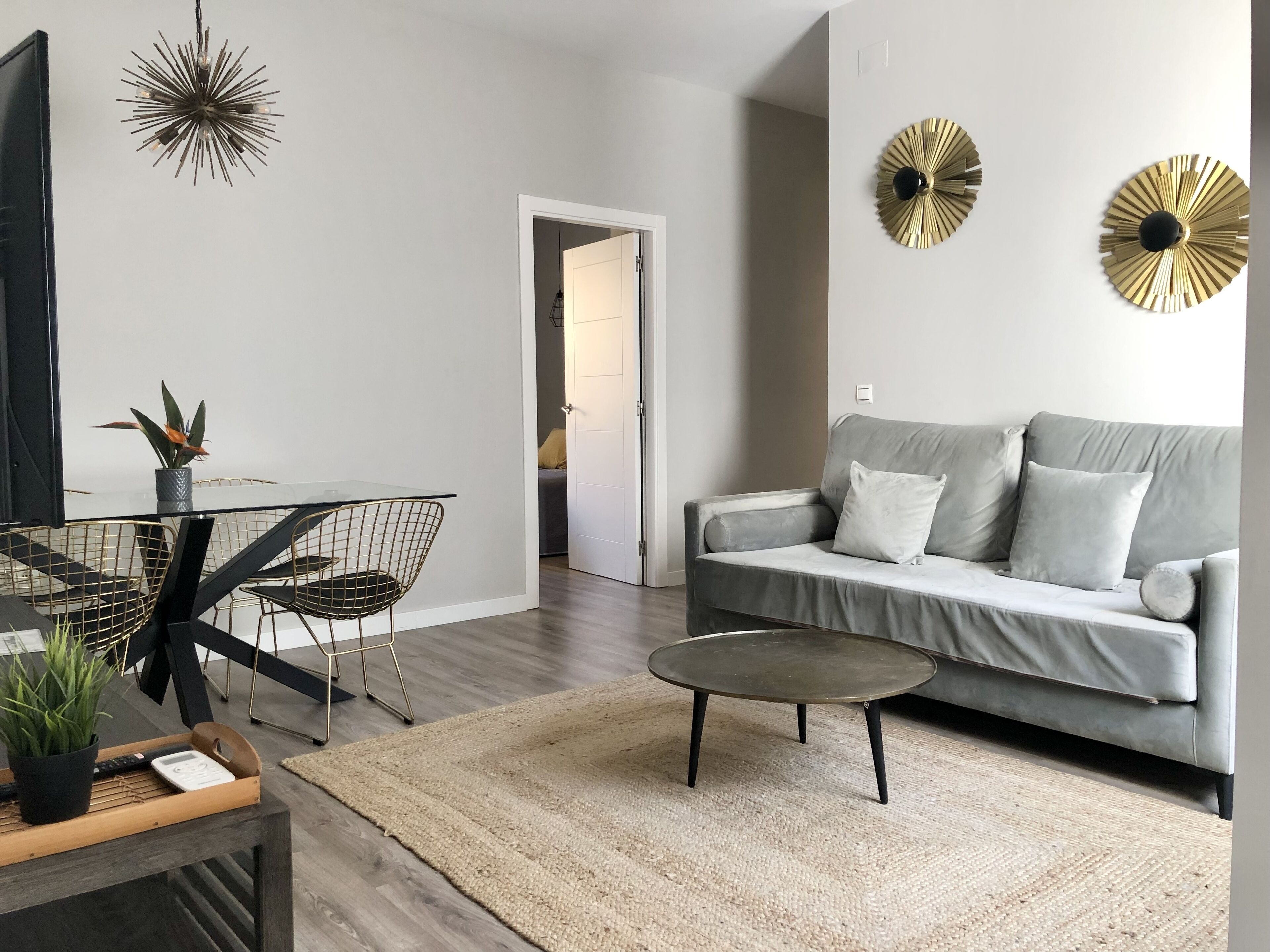 A modern living room with a grey sofa, a coffee table, a dining area with a glass table and wire chairs, and decorative wall art.