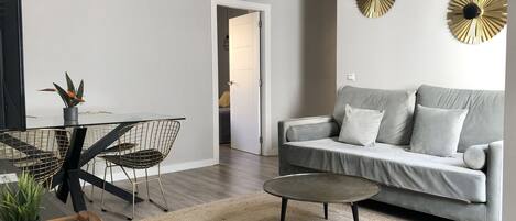 A modern living room with a grey sofa, a coffee table, a dining area with a glass table and wire chairs, and decorative wall art.