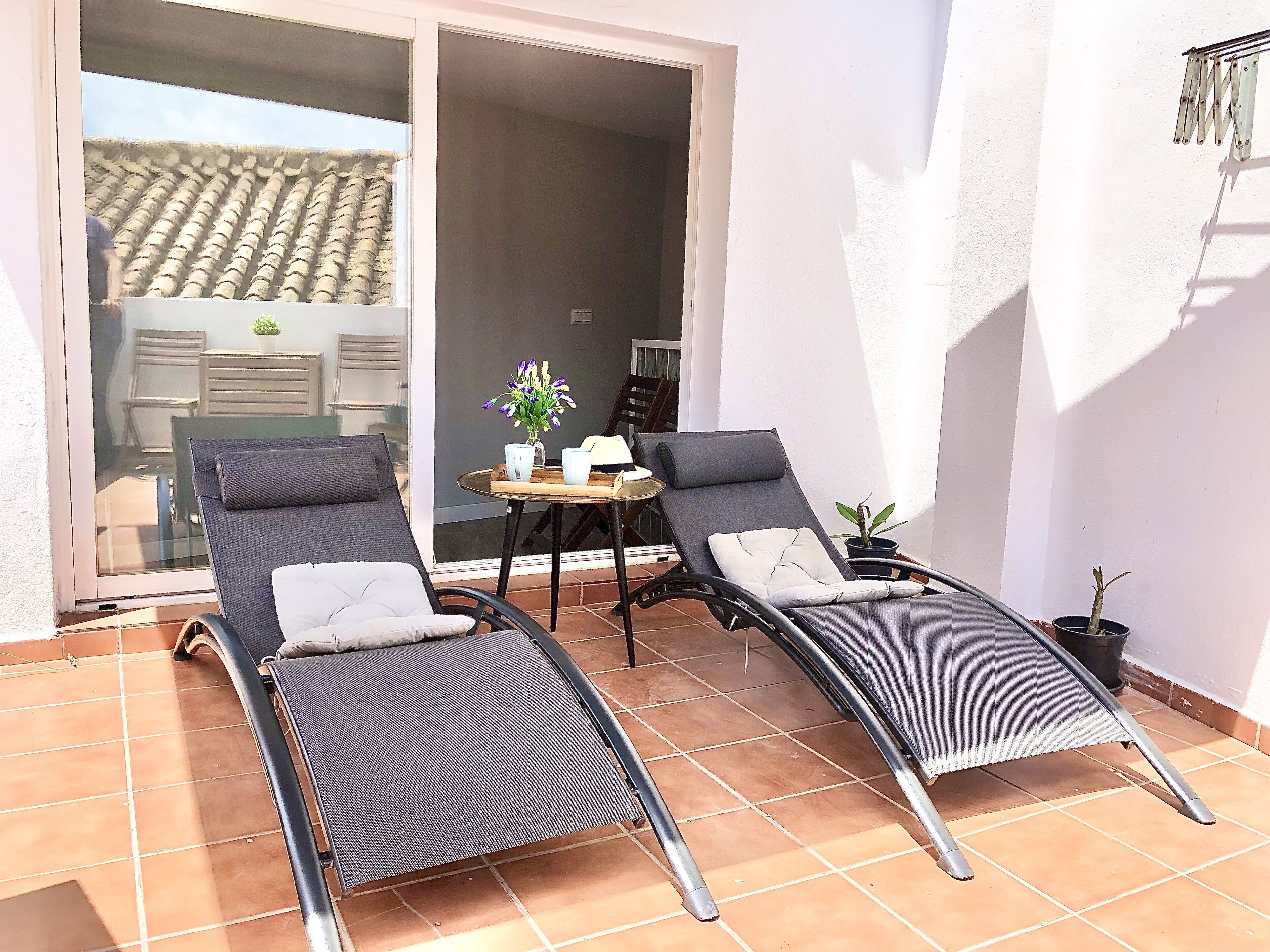 A sunlit patio with two reclining chairs, a small table with a vase of flowers, and a sliding glass door leading to an indoor area.