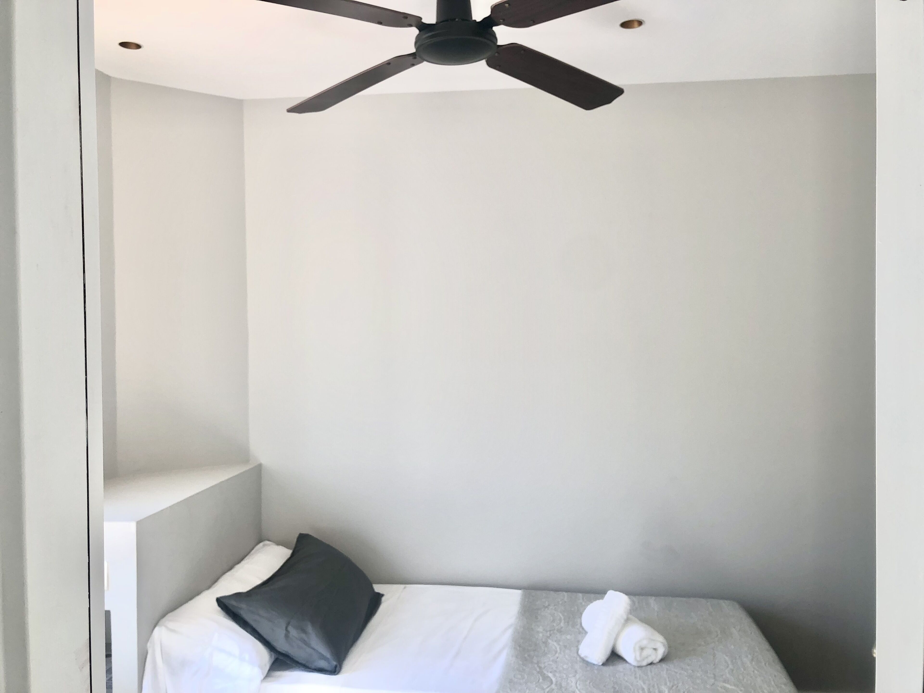 Una habitación pequeña con cama individual, ventilador de techo, paredes blancas y un colchón gris texturizado.