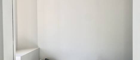 Una habitación pequeña con cama individual, ventilador de techo, paredes blancas y un colchón gris texturizado.