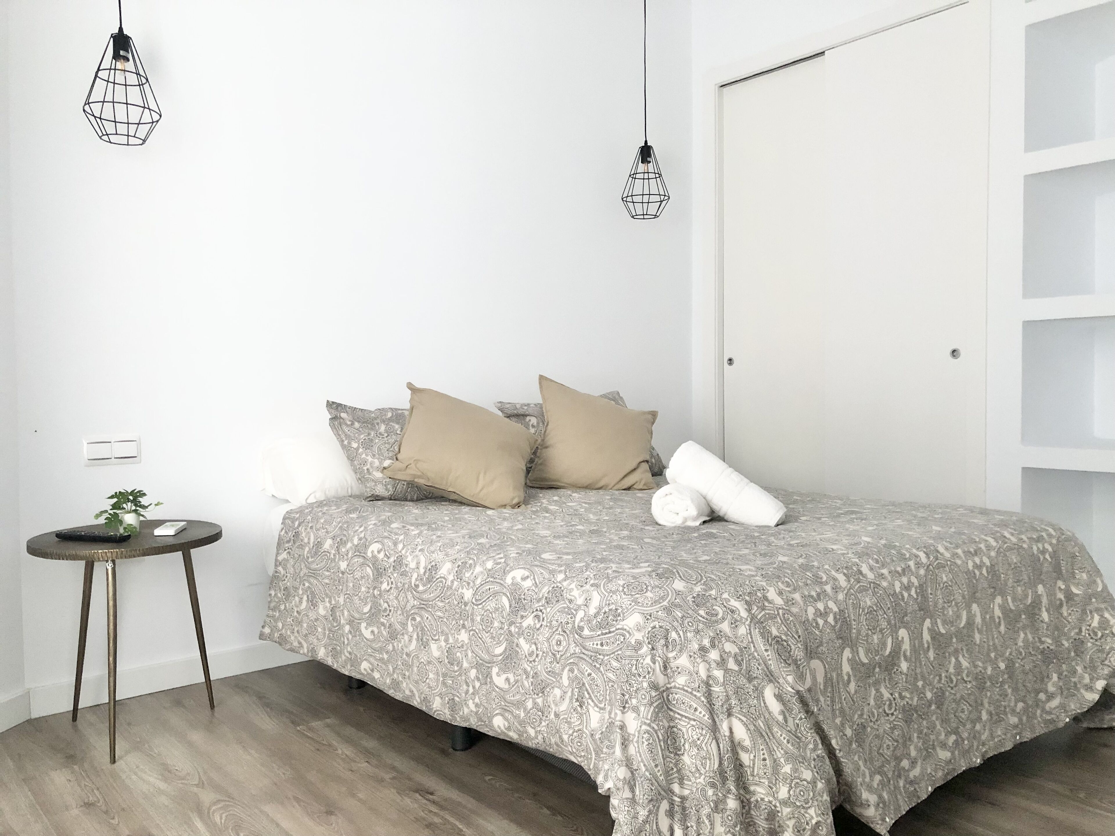 A bedroom with a bed, two hanging light fixtures, a small round table with a plant, and a white door.