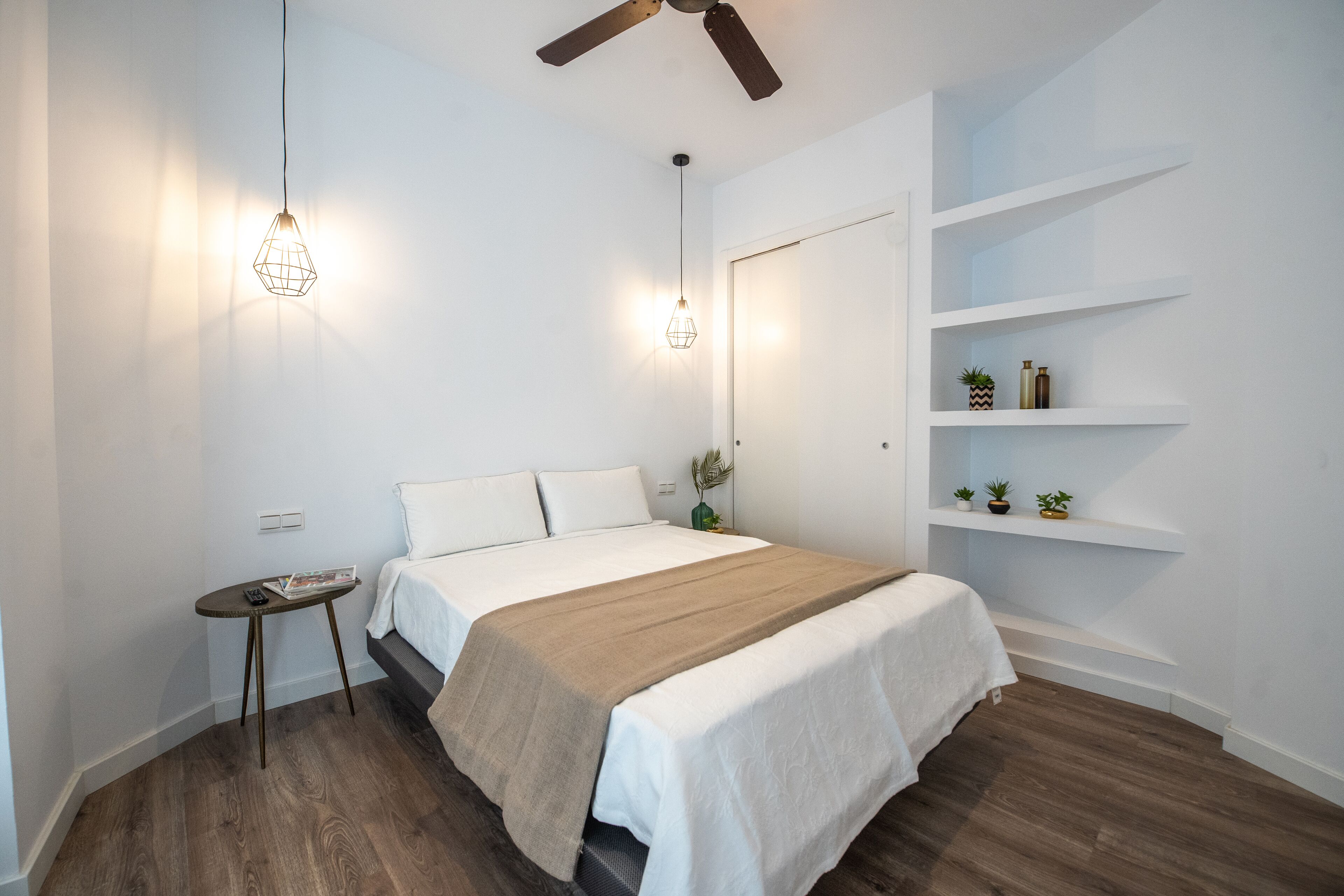 A modern bedroom with a bed, bedside tables, a ceiling fan, and built-in shelving with decorative items.