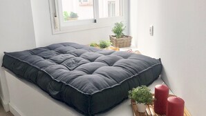 Una cama pequeña y ordenada con un colchón oscuro y almohadas, una ventana con persianas, una mesita con una planta y velas, y una cesta de mimbre con más plantas.