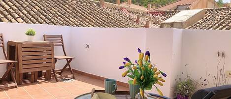 Una terraza con techo de tejas, una mesa con sillas, un jarrón con flores y vistas a una colina con edificios.