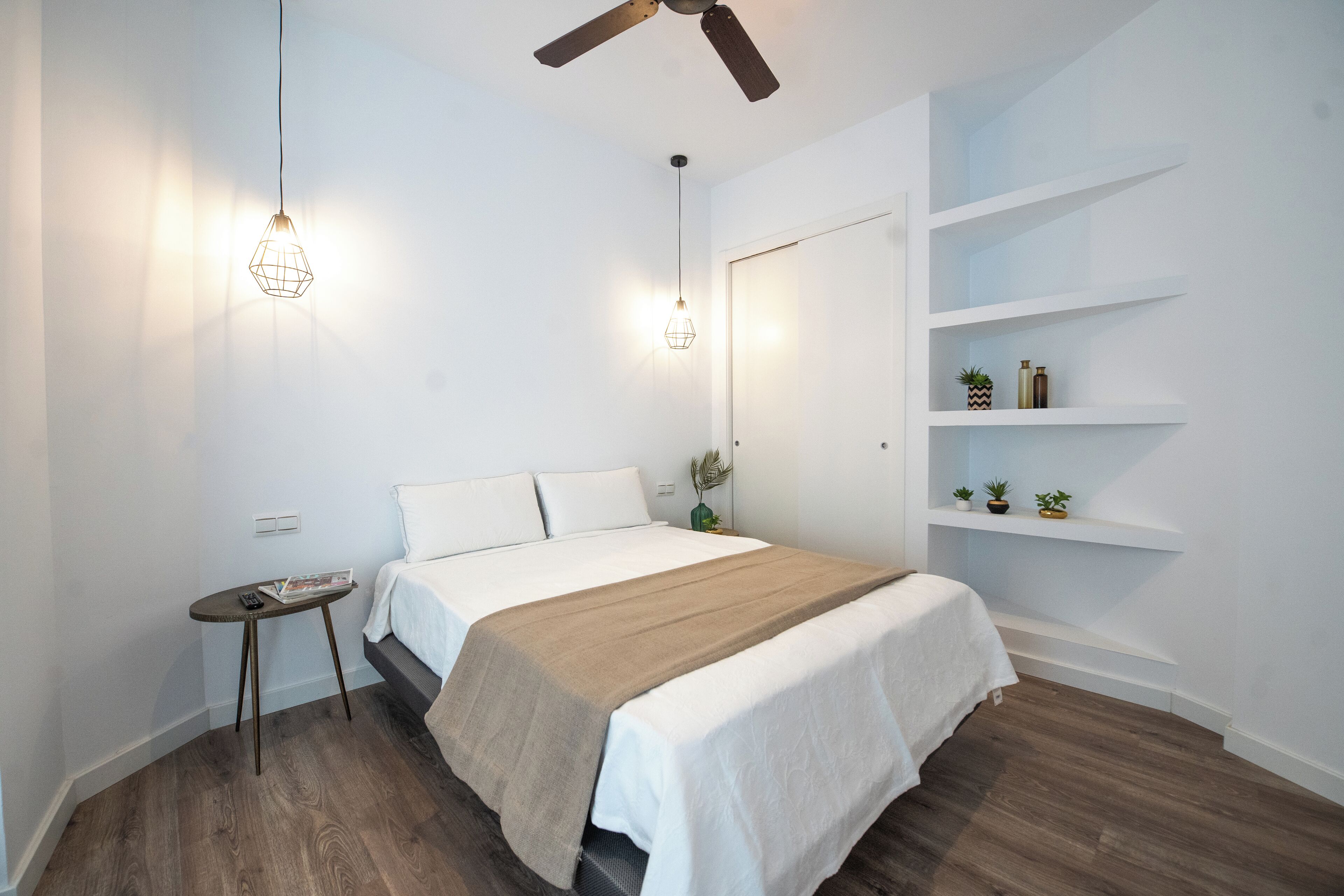 A modern bedroom with a bed, bedside tables, a ceiling fan, and built-in shelving with plants.