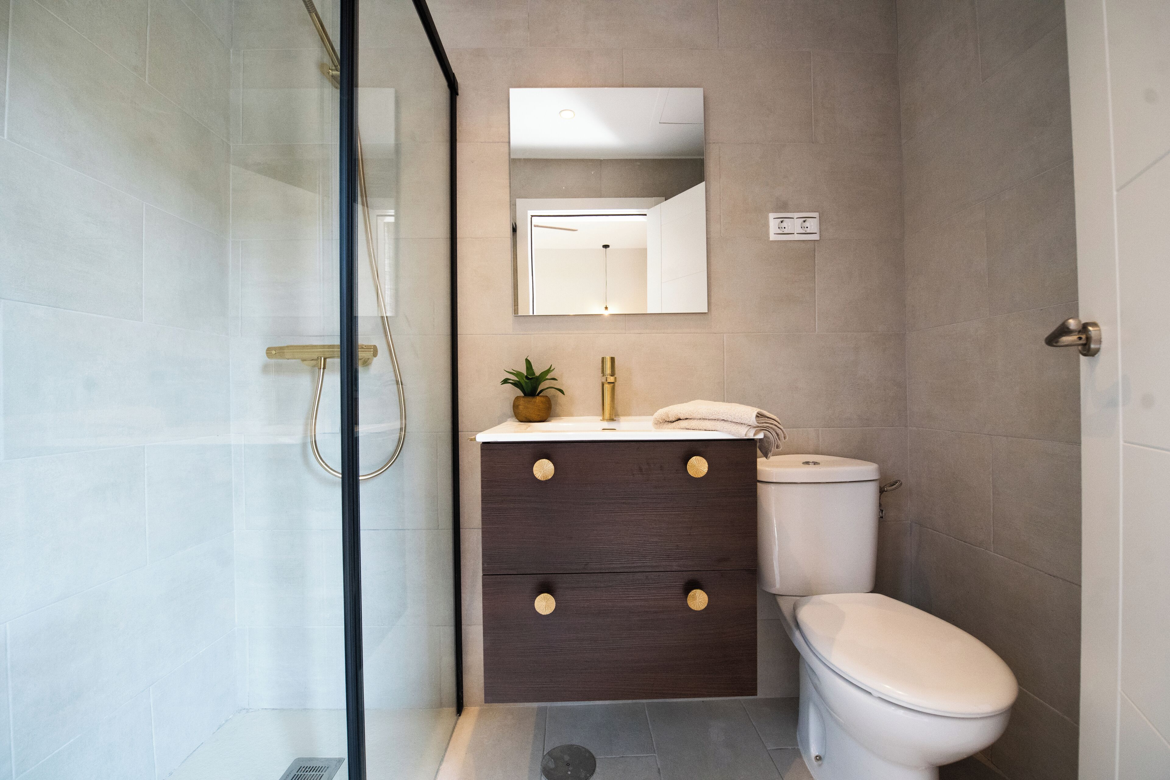 A modern bathroom with a glass shower enclosure, a dark wooden vanity with gold handles, a white toilet, and a mirror above the vanity.
