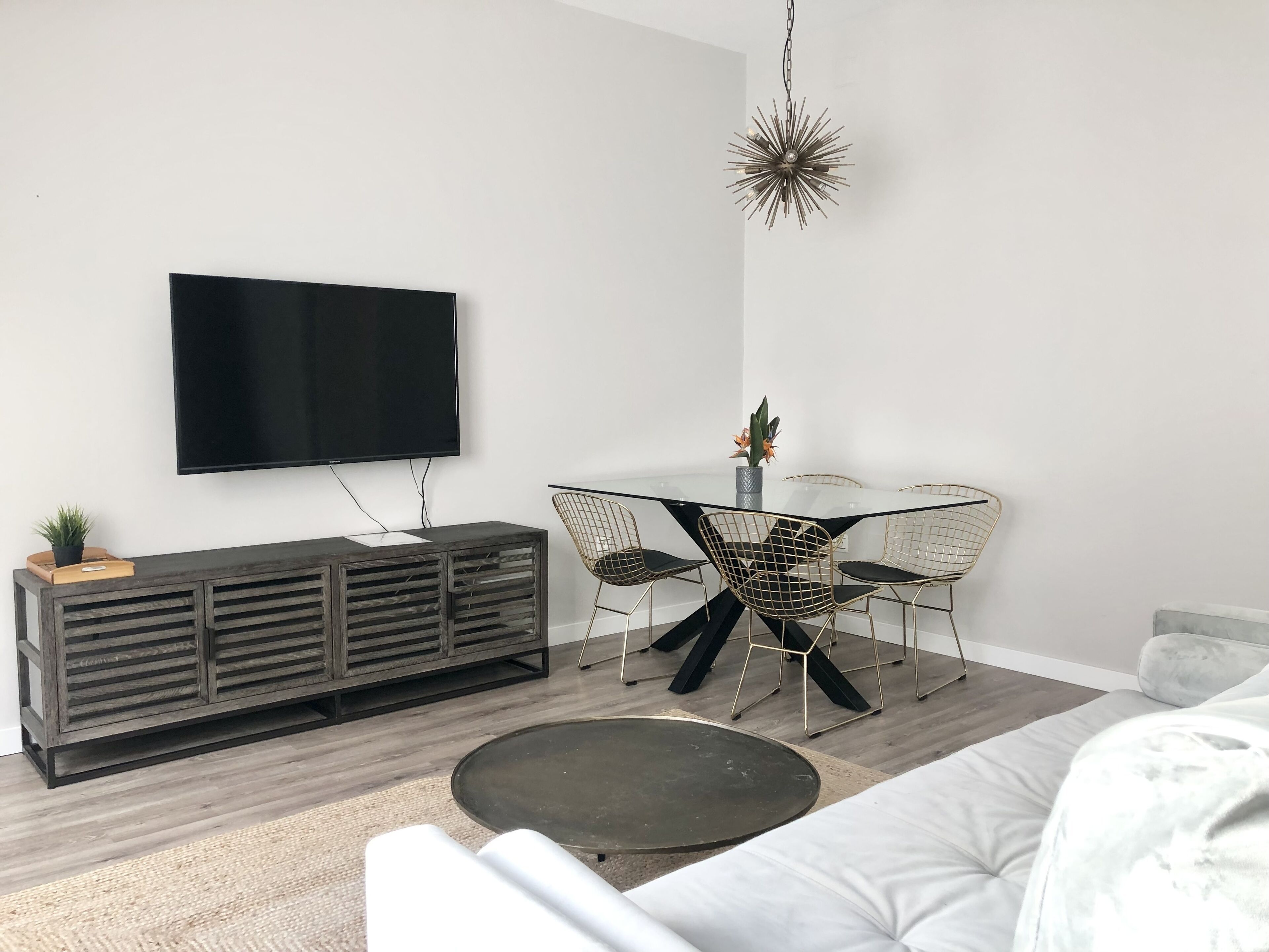 A modern living room with a flat-screen TV, a round glass table, wire mesh chairs, a dark wooden console, and a grey sofa.