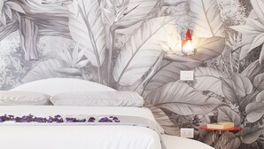 Een slaapkamer met een wit bed, een nachtkastje met een rode lamp en een muurschildering van tropische bladeren.