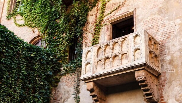 A historic building with a balcony featuring intricate stonework and a brick facade partially covered by green ivy. - Αn enchanting residence in front of the Juliet house (Verona)