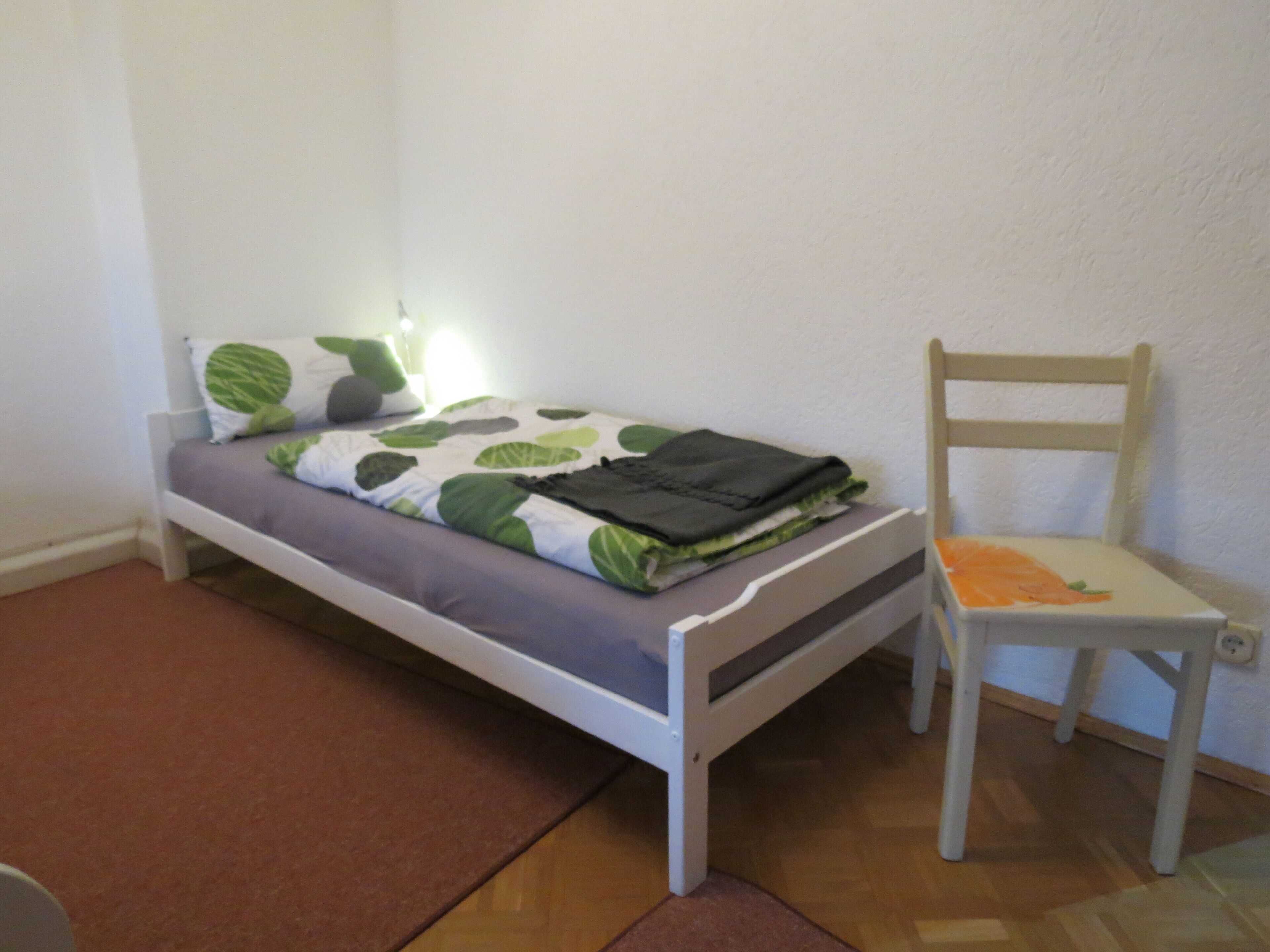 Una habitación pequeña con una cama individual, una silla de madera y una mesita de noche.