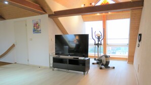 Un vaste salon doté d’un grand téléviseur à écran plat posé sur un meuble noir, d’un vélo d’appartement et d’un escalier menant à un niveau supérieur.