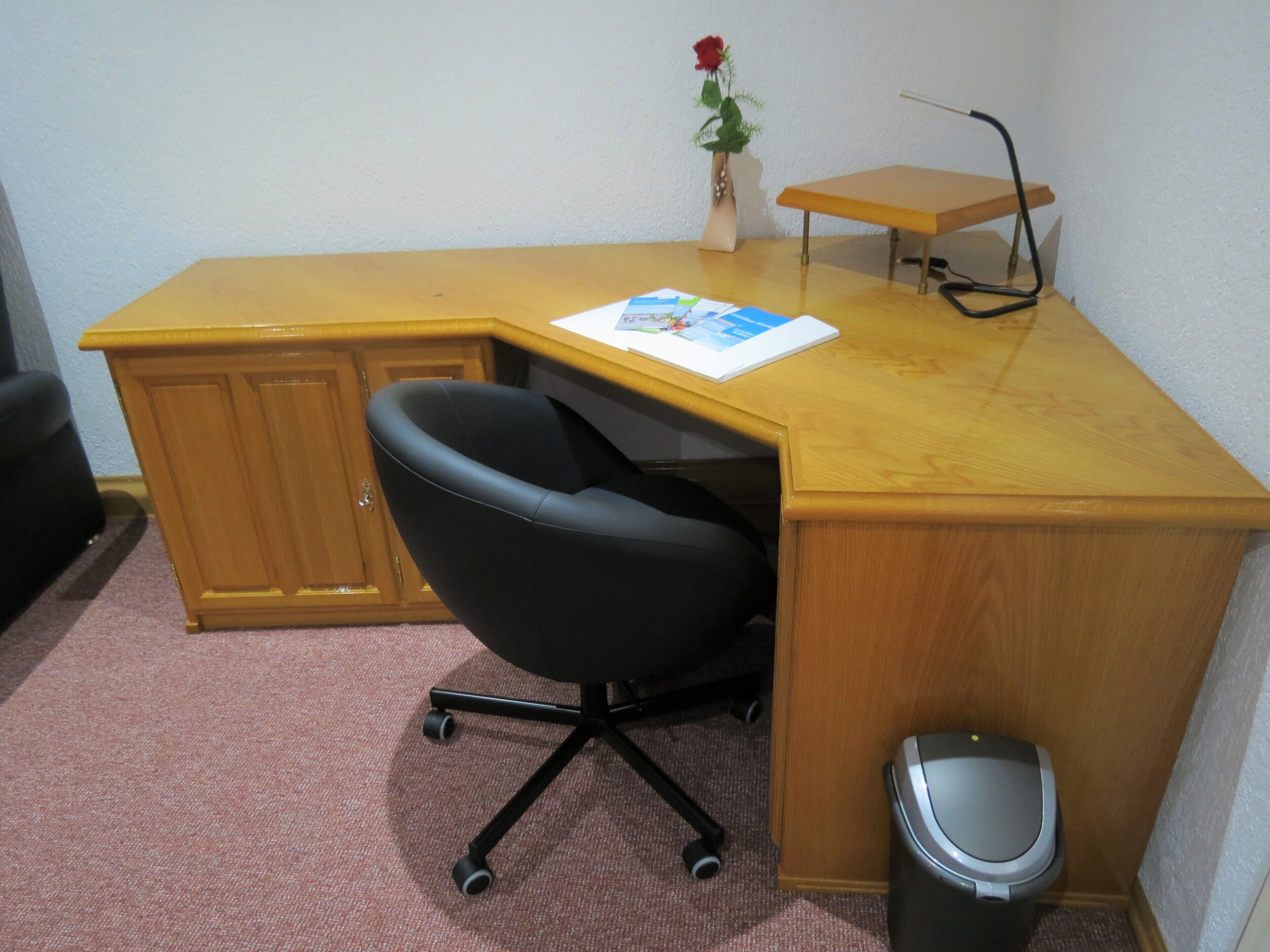 Un petit bureau avec un bureau en bois, une chaise de bureau noire, une corbeille à papier et un petit vase contenant une seule rose rouge.