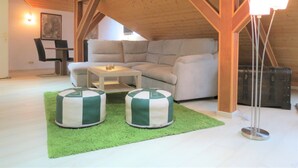 Une pièce mansardée chaleureuse, avec un canapé d’angle, une petite table, deux fauteuils en forme de poire, un lampadaire et un coin repas comprenant des chaises et une table.