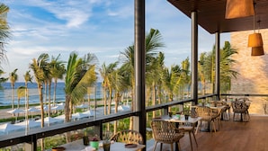 A terrace with tables and chairs, overlooking a beach with palm trees and the ocean.