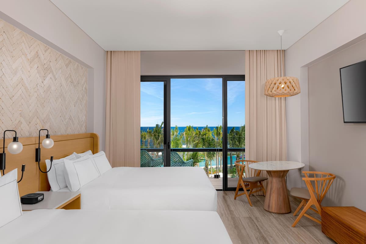 A hotel room with a large bed, a desk, a chair, a small table, and a TV. The room has a large window with a view of the ocean and palm trees.