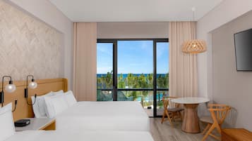 Habitación de hotel con una cama grande, un escritorio, una silla, una mesita y un televisor. La habitación tiene un ventanal con vistas al océano y palmeras.