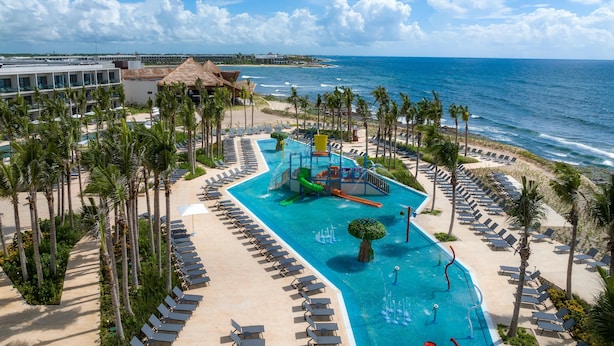 A resort pool with a water slide, surrounded by palm trees and lounge chairs, overlooking the ocean.
