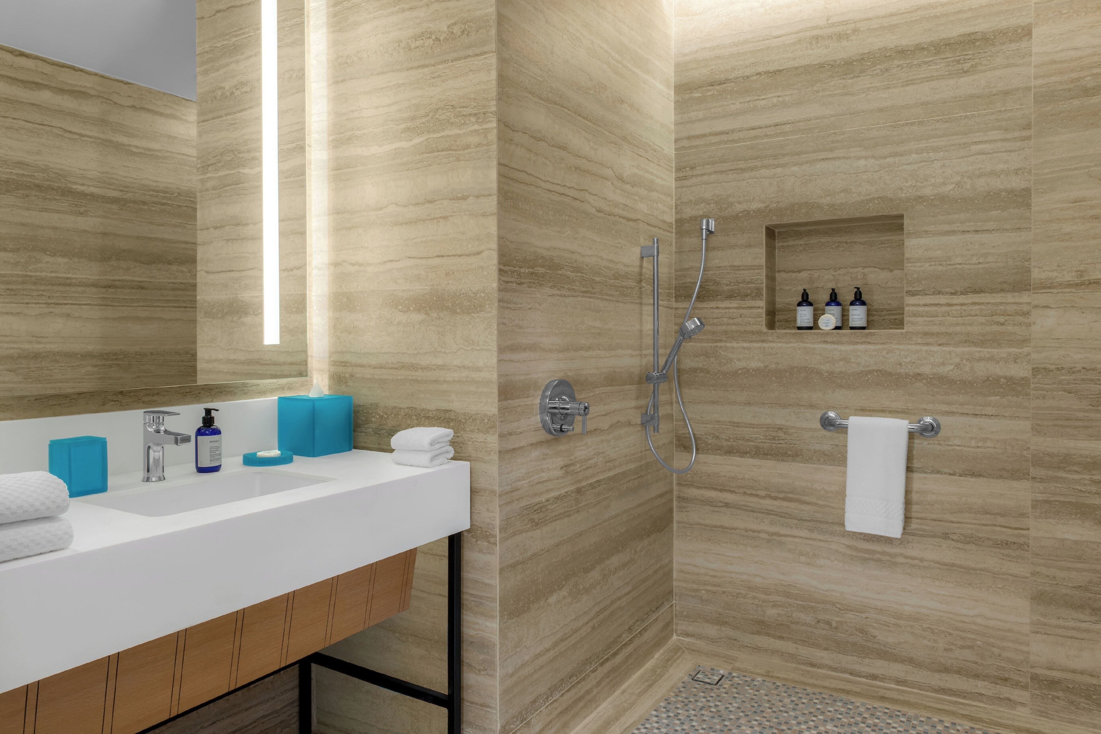 Une salle de bain moderne avec un lavabo blanc, des distributeurs de savon bleus, une cabine de douche en verre et un porte-serviettes.
