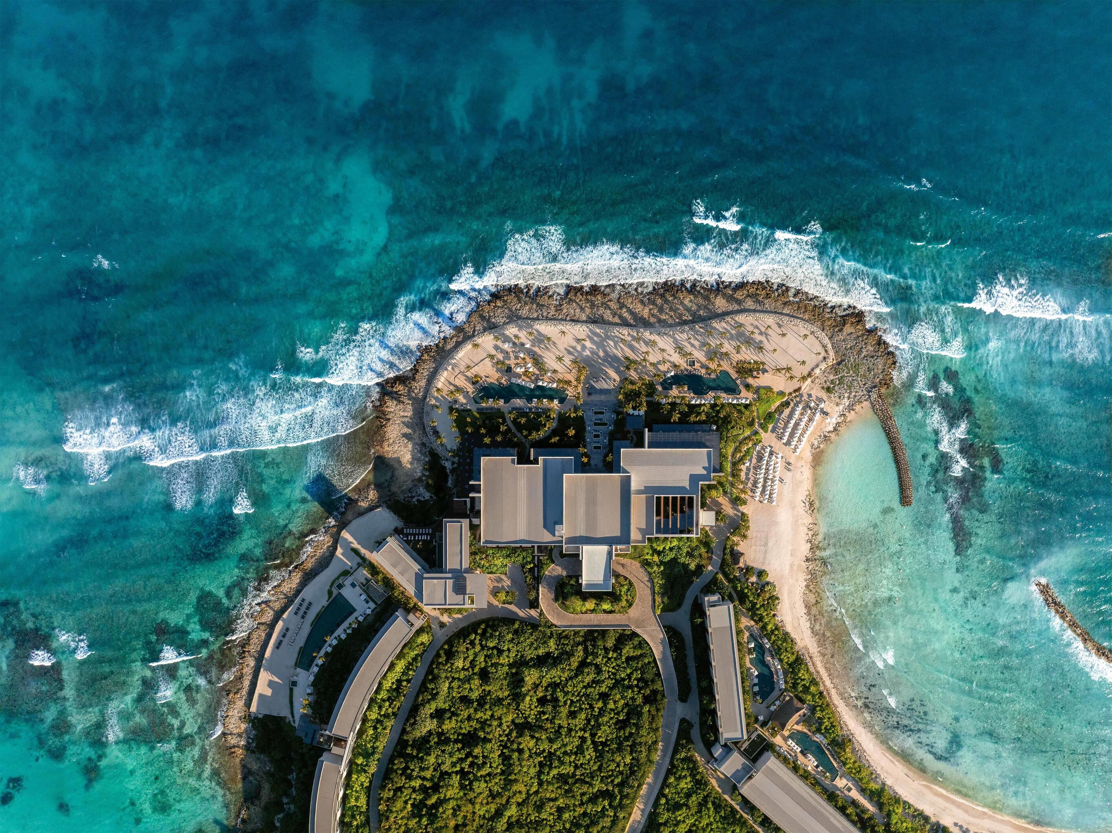 An aerial view of a coastal resort with a central building surrounded by a beach and a pool area.