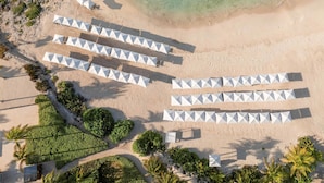 An aerial view of a beach with rows of white umbrellas and lounge chairs, surrounded by greenery and a clear blue sea.