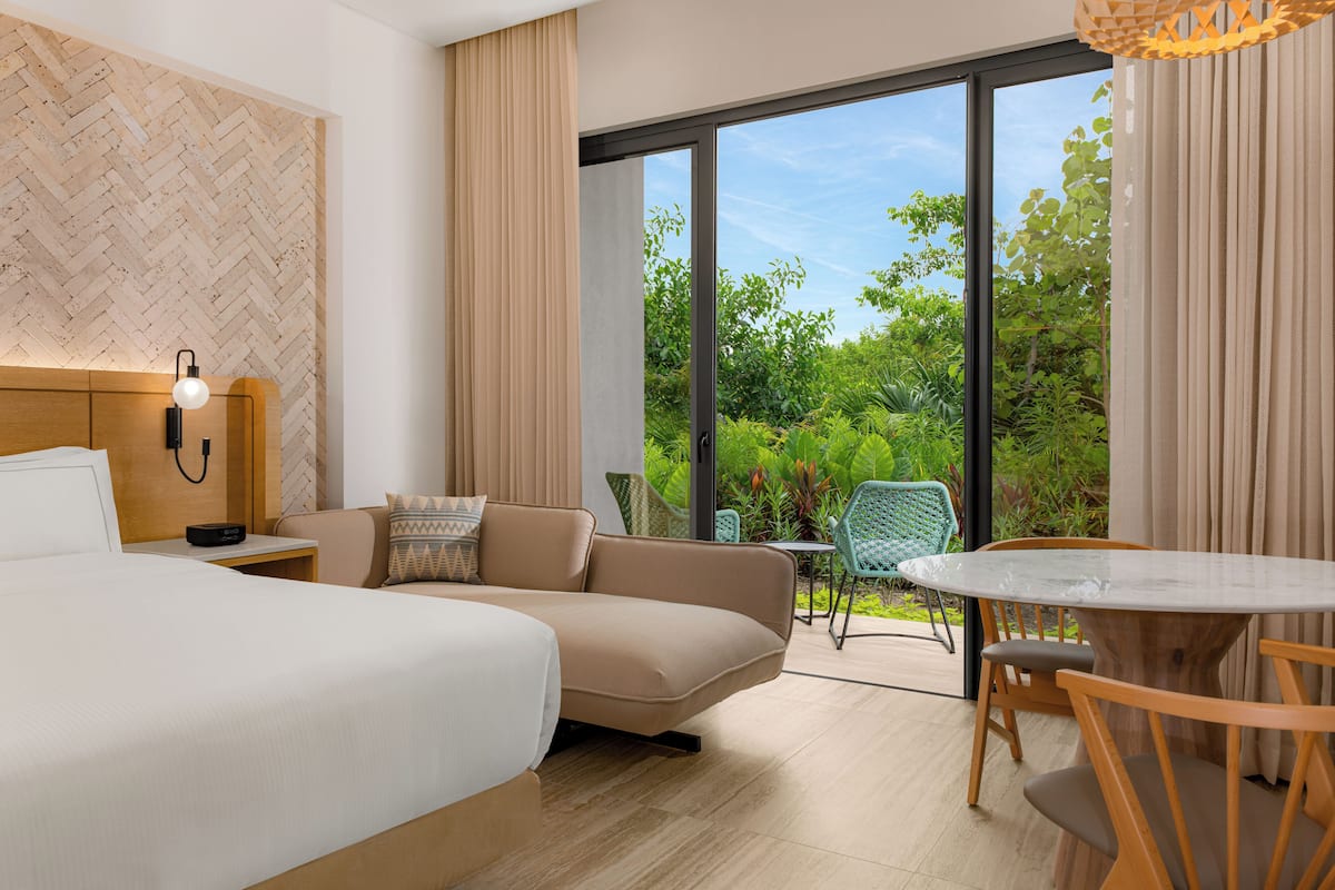 A modern hotel room with a bed, a sofa, a dining table, and a view of greenery through large windows.