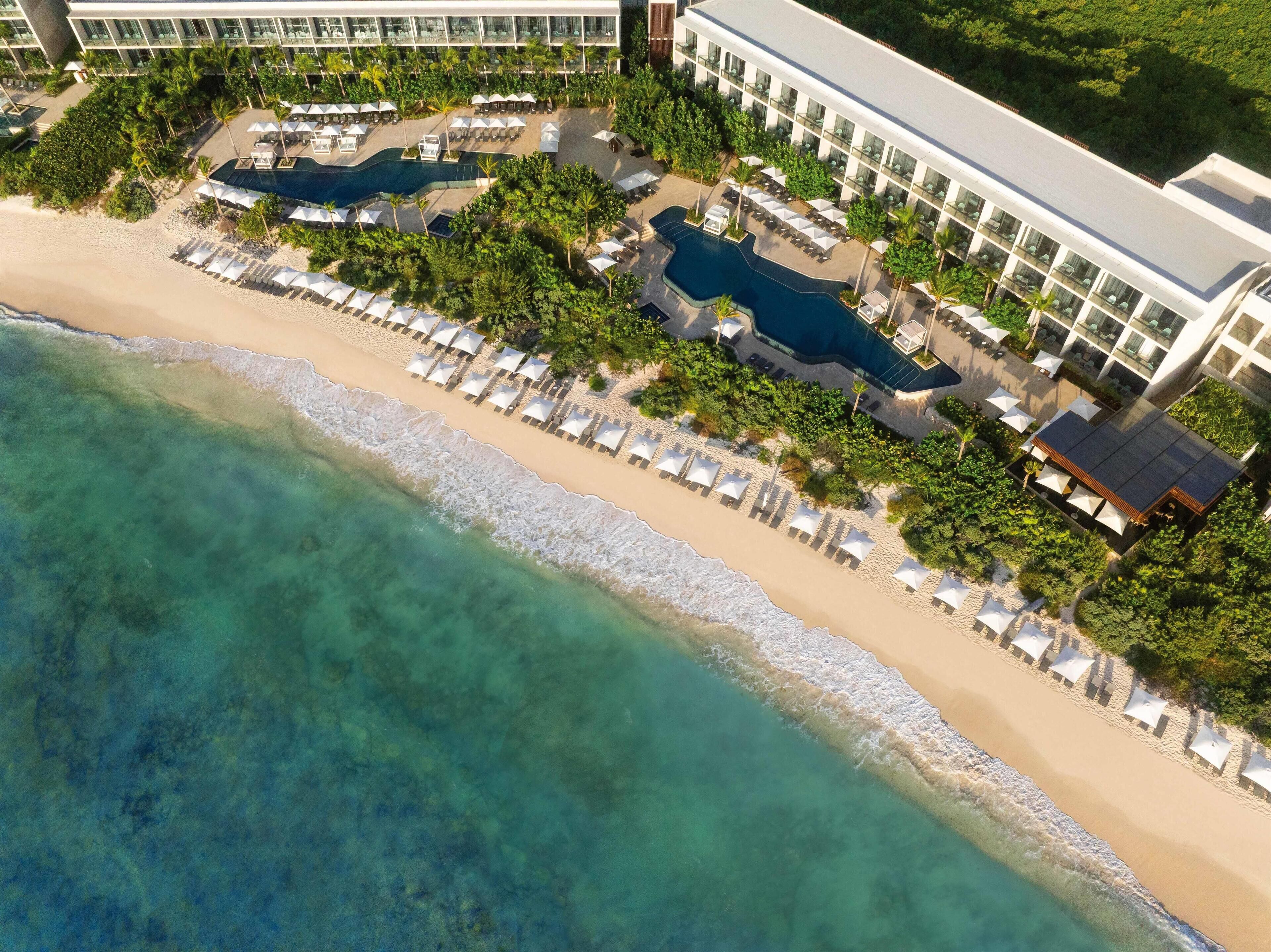 An aerial view of a resort with a beach, a pool area, and a building with multiple balconies.