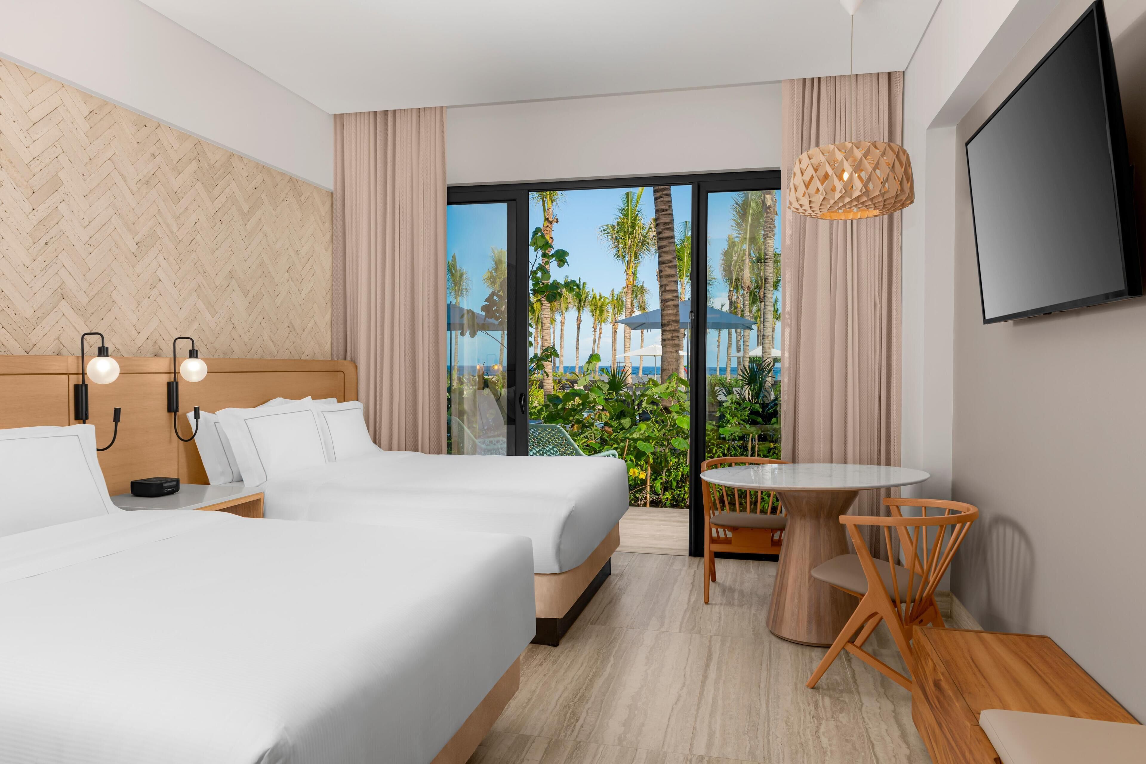 A hotel room with two beds, a TV, a small table with chairs, and a view of palm trees and the sea.